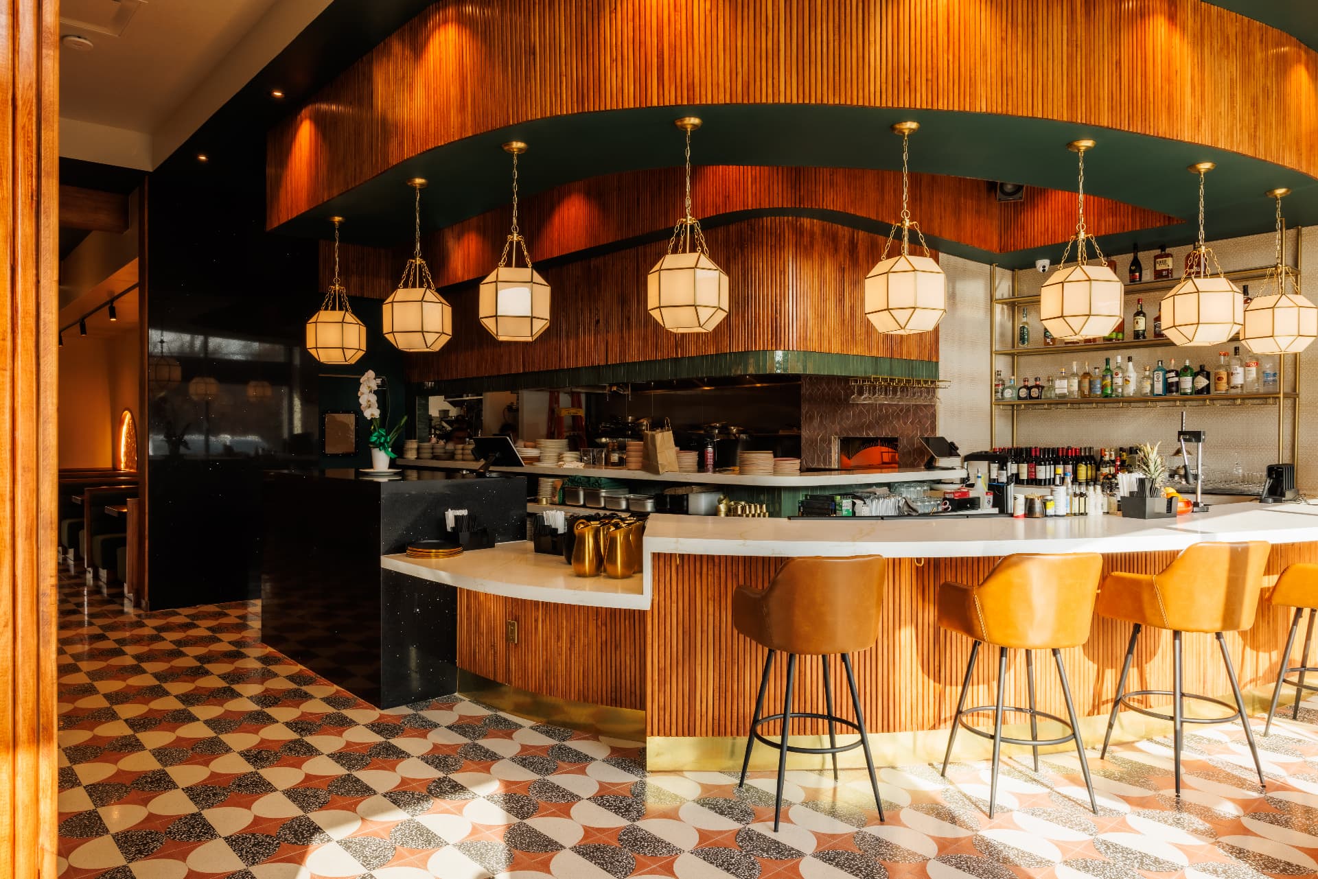Sunlit restaurant bar with fluted wood paneling, cognac leather stools, brass shelving, and geometric lantern pendants
