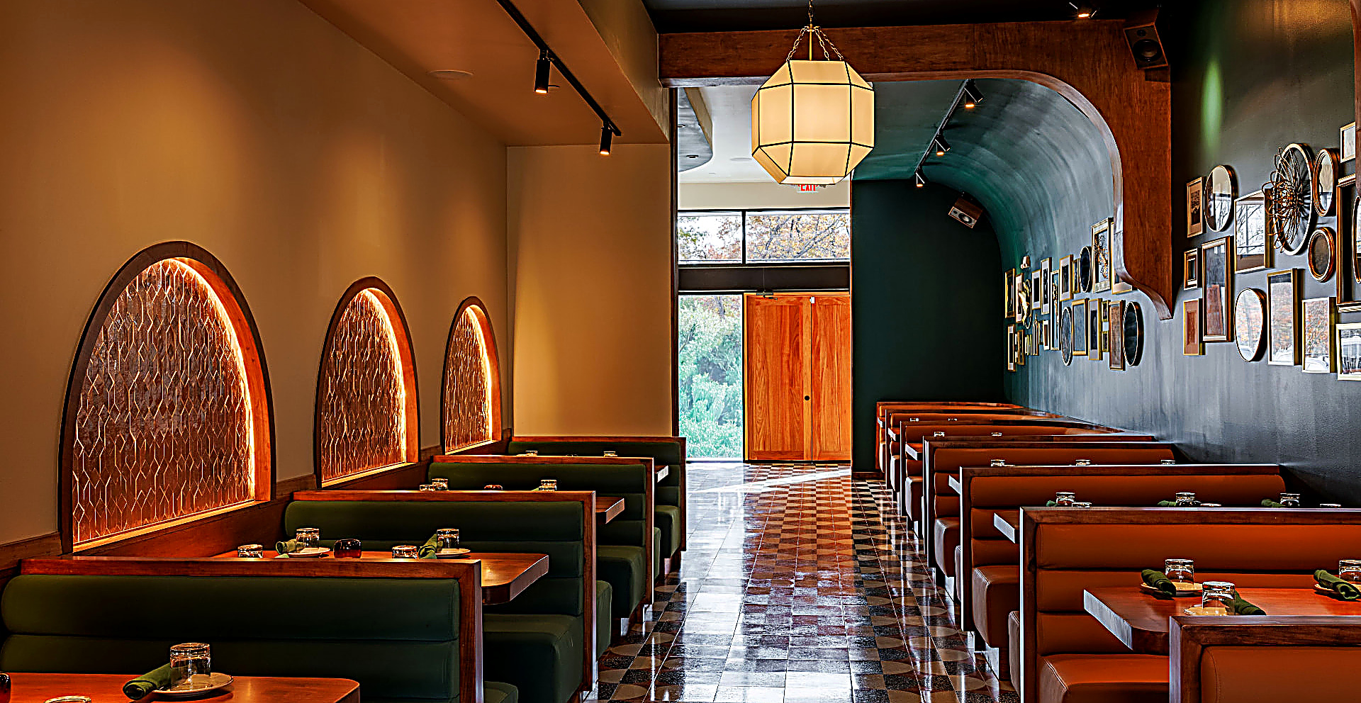 Zon Zon dining room — a patterned tile aisle running toward sunlit double doors, backlit arched niches above green booths on the left, cognac leather booths beneath a gallery wall of framed art on the right, cream pendant lanterns overhead