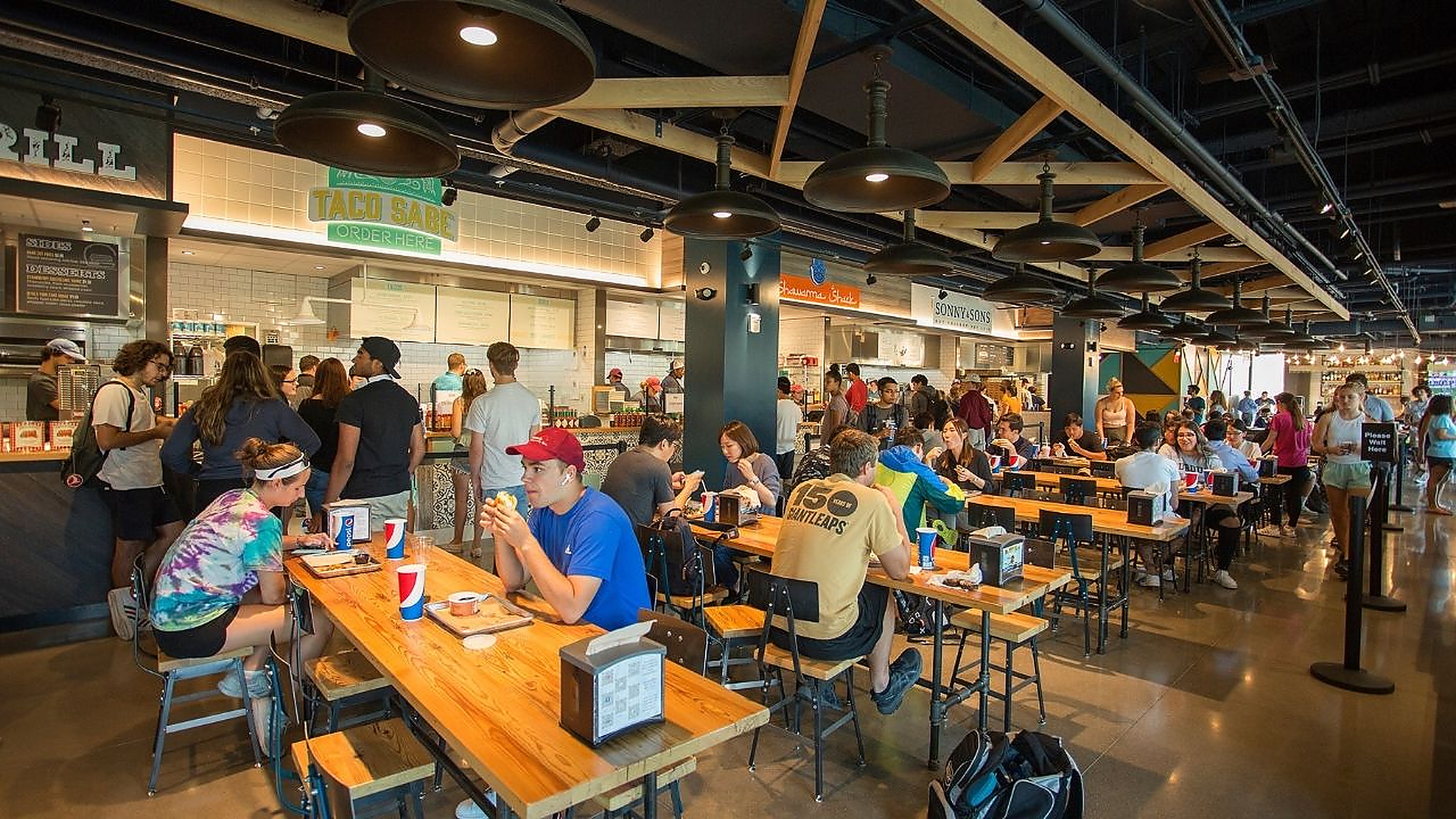 The main hall at Crave Food Hall at Purdue — students at long blonde-wood communal tables beneath timber rafters and rows of matte black dome pendants, with the Taco Sabe, Shawarma Shack and Sonny & Sons stalls along the back wall