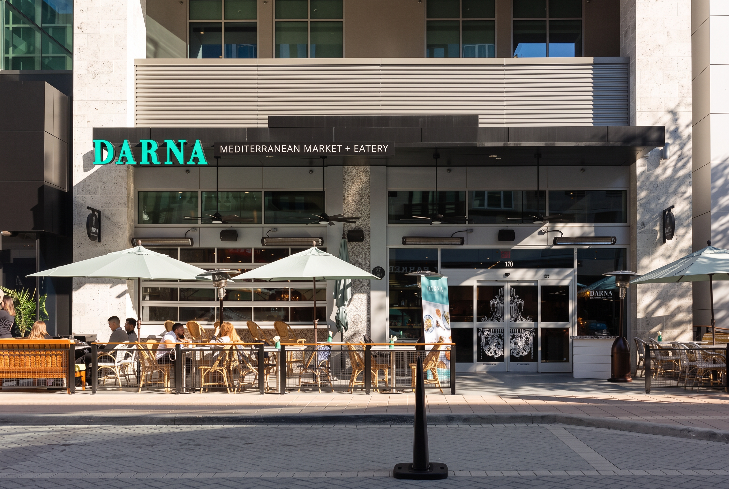 Darna Mediterranean Market + Eatery storefront with turquoise signage, rattan patio seating, and canvas umbrellas on a sunlit sidewalk