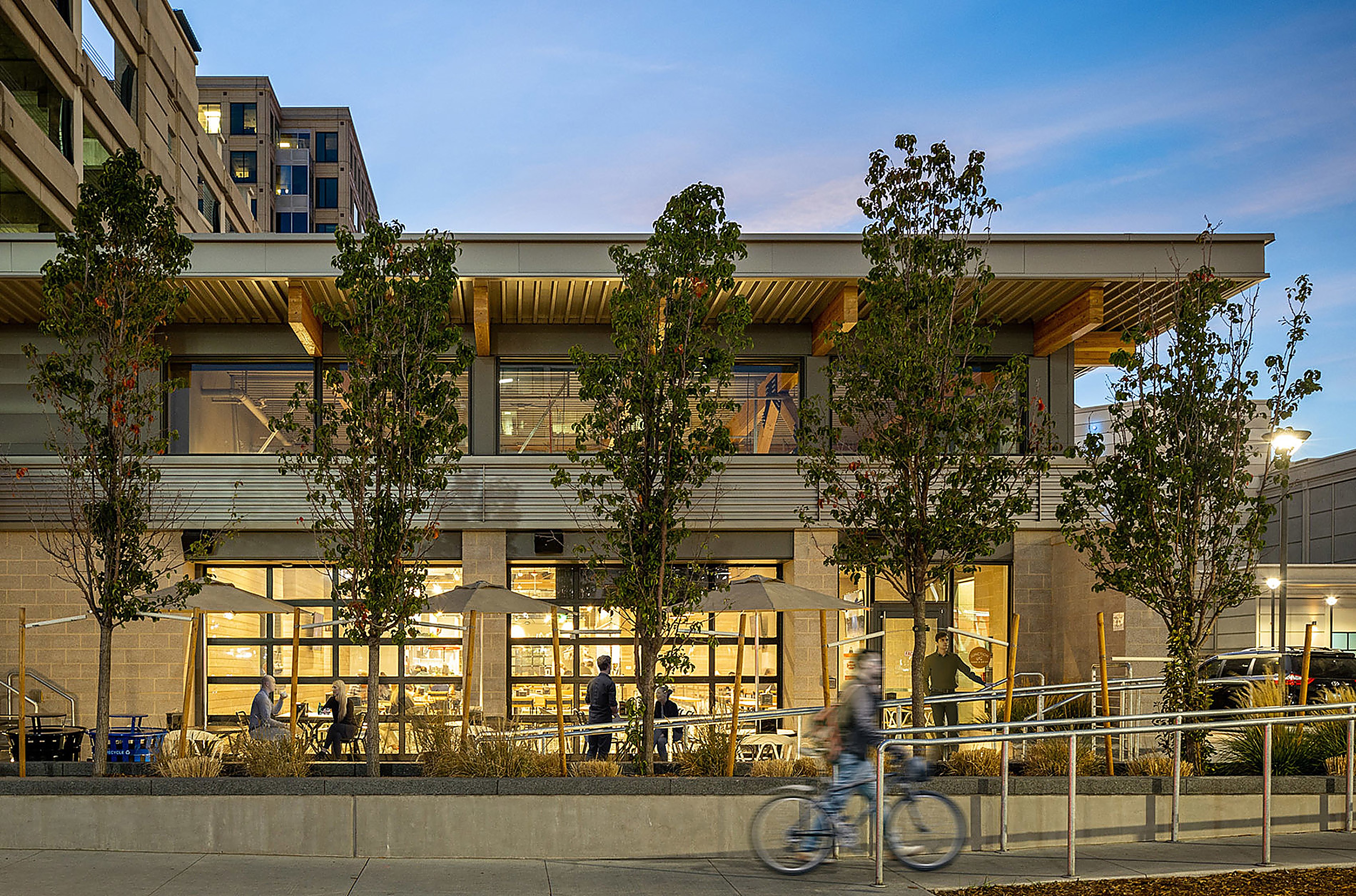 The Junction exterior at twilight — a two-storey building with roll-up glass garage doors glowing warm from inside, an outdoor patio with umbrellas along the sidewalk, and a cyclist blurred across the foreground